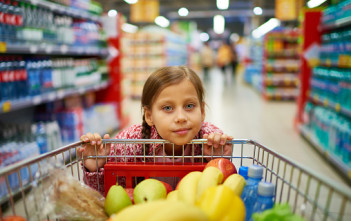 Cute girl leaning on trolley with products while shopping in supermarket and looking at camera