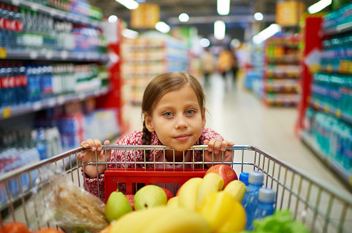 Cute girl leaning on trolley with products while shopping in supermarket and looking at camera