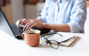 Remote working from home. Freelancer workplace in kitchen with laptop, cup of coffee, spectacles. Concept of distance learning, isolation, female business, shopping online. Close up of woman hands.