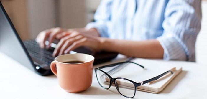 Remote working from home. Freelancer workplace in kitchen with laptop, cup of coffee, spectacles. Concept of distance learning, isolation, female business, shopping online. Close up of woman hands.