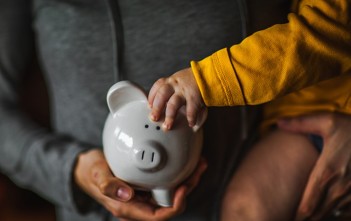 A baby holds on to the ear of a piggy bank while being held by its mother.