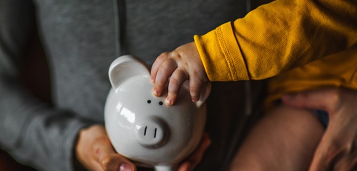 A baby holds on to the ear of a piggy bank while being held by its mother.